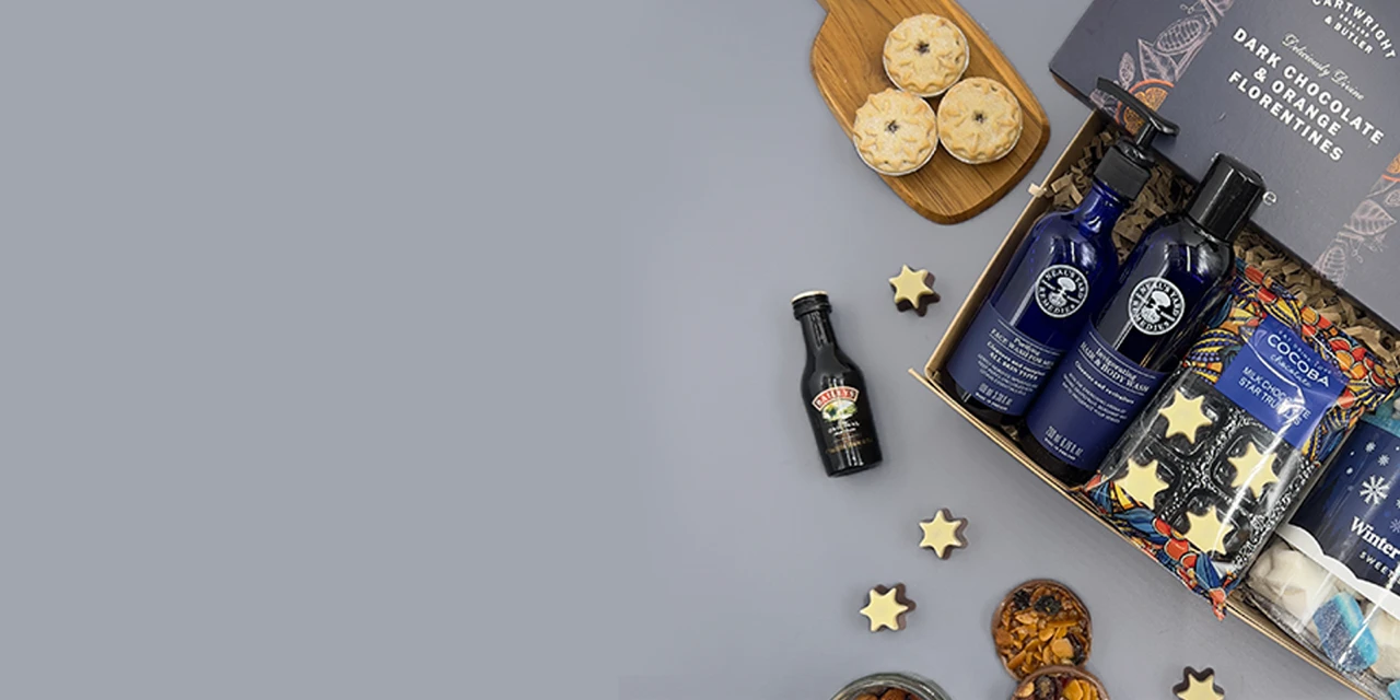 Luxury Christmas hamper for him with artisan chocolate, festive snacks, premium bottles, hot chocolate and gourmet treats arranged in a gift box on a grey background.