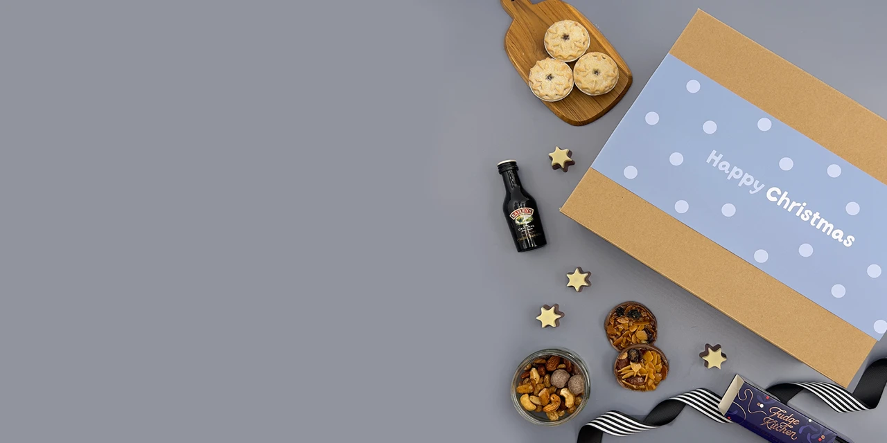 Luxury Christmas hamper for him with artisan chocolate, festive snacks, premium bottles, hot chocolate and gourmet treats arranged in a gift box on a grey background.