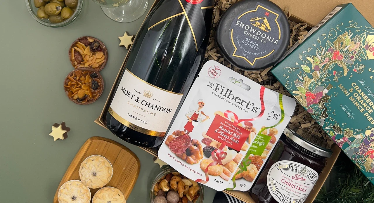 Luxury Christmas food hamper with champagne, gourmet snacks, olives, artisan crackers and festive treats beautifully arranged on a soft sage green background.