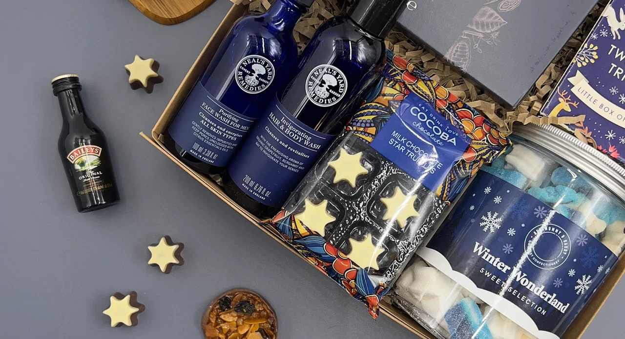 Luxury Christmas hamper for him with artisan chocolate, festive snacks, premium bottles, hot chocolate and gourmet treats arranged in a gift box on a grey background.